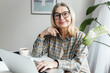 © shurkin_son - Happy smiling elderly lady in casual clothes and glasses watching movie or series online on laptop using web application, pressing pause while looking at camera, sitting at table with cup of tea