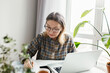 © shurkin_son - Retired people and remote education. Upper angle indoor view of intelligent senior caucasian woman in glasses writing brief summary in copybook while study online at home using laptop
