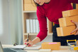 © ThawKyar - Asian female small business owner working from home taking orders and shipping to customers with laptop, pen and notepad. Ecommerce concept for SMEs.