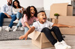 © Prostock-studio - At New Apartment. Excited African American family having fun and celebrating moving day, cheerful girl riding her brother in cardboard box, parents emracing sitting on the sofa in blurred background