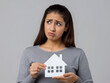 © Hasanka - Young Indian woman with ponytail, a real estate agent, holds a white house-shaped cardboard cutout. She looks at a house model with a worried expression, set against a light grey background.
