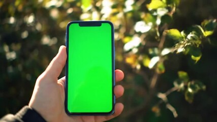First-person shot holding a green screen phone outdoors. Ideal for app promotion, social media content, and urban mobile usage mockups.