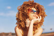 © SHOTPRIME STUDIO - Curly haired woman wearing stylish sunglasses, posing with a thoughtful expression against a bright blue sky