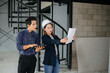 © laddawan - Asian engineers wearing hard hats discuss blueprints and floor plans at a construction site. Teamwork, planning, and innovation