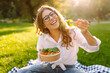 © maxbelchenko - Happy woman in glasses eating salad on a picnic mat. Young woman enjoying a healthy bowl and basking in the sun in a sunny park. Concept of relaxation, enjoyment.