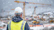© Maksym - Wide shot of construction worker in white helmet surveying snow-covered site, footprints in fresh snow, large cranes and bulldozers parked under grey winter sky, muted cold tones e