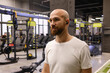 © arthurhidden - Man standing in gym with fitness equipment, wearing wireless earbuds and white t-shirt