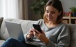 © li - Woman using laptop and typing on a phone while sitting on couch at home. High quality