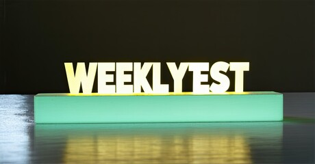 Poster - Bright illuminated word WEEKLYEST rests upon turquoise rectangular base. Reflections visible on glossy surface below. Simple studio setup.