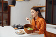 © SHOTPRIME STUDIO - Young woman enjoying a healthy salad in a cozy kitchen, wearing an orange shirt, displaying happiness and relaxation, with a focus on healthy eating
