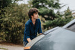 © qunica.com - A young man stands next to his vehicle, inspecting it intently on a rainy day. The scene captures a moment of concentration and concern in an outdoor setting.