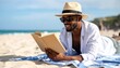 © David Andres - Un hombre joven con sombrero de paja y gafas de sol tumbado sobre una toalla en una playa y leyendo un libro. Vacaciones y relax