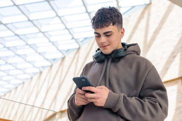 young man using smartphone with headphones around neck in modern glass building, teenager texting on