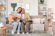 © Pixel-Shot - Happy young woman playing guitar with her grandmother on sofa at home