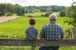© Vlad - Father Son Farm. Back View of Father and Son Sitting on Fence, Admiring Beautiful Green Summer Field