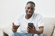 © SHOTPRIME STUDIO - Smiling Black man in a casual white t shirt using a smartphone, showcasing engagement and happiness in a bright, minimalistic living room setting