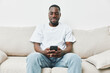 © SHOTPRIME STUDIO - Young Black man sitting on a beige couch, smiling while using a smartphone, showcasing casual fashion and relaxed emotions in a modern living space