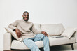 © SHOTPRIME STUDIO - Happy young Black man sitting on a sofa in cozy sweater, smiling and enjoying a relaxed moment at home, promoting comfort and lifestyle concepts