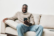 © SHOTPRIME STUDIO - Smiling Black man in cozy sweater using smartphone on sofa in a modern living room, showcasing relaxation and connection in a contemporary space