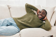 © SHOTPRIME STUDIO - Happy black man relaxing on a sofa in a cozy living room, wearing a green sweater and jeans, conveying comfort and joy in a modern, bright space