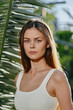 © SHOTPRIME STUDIO - Young woman with long brown hair wearing white tank top standing near palm leaves in soft natural sunlight outdoors, showcasing serene expression and casual style.