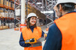 © Koldo_Studio - Smiling female warehouse worker holding handheld computer and clipboard discussing logistics with colleagues in large distribution center
