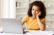 © Anastasiya - Unhappy African American woman student sitting at desk and looking at laptop, upset black female leaning on hand, feeling tired or bored during online lesson, closeup portrait