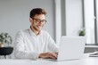 © ttonaorh - A handsome man in a shirt and glasses is sitting at a white table with a laptop, smiling while working on a digital marketing project in a modern office room with copy space.
