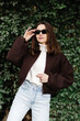 © nazariykarkhut - Stylish woman adjusting sunglasses while posing near ivy wall