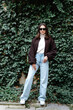 © nazariykarkhut - Stylish woman posing in front of green ivy wall wearing sunglasses, turtleneck sweater and bomber jacket