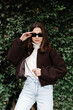 © nazariykarkhut - Stylish woman adjusting sunglasses while posing near ivy wall