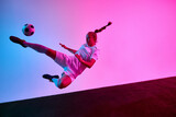 Dynamic photo. Young female footballer performing flying side kick in air with braided hair and full focus under pink blue neon light. Concept of sport, agility, performance, athletic excellence.