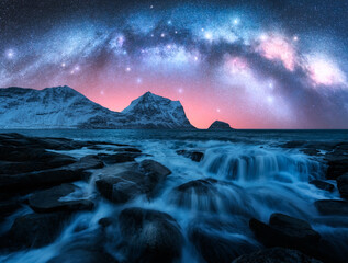 Wall Mural - Night starry sky with glowing Milky Way, rocky shore with stones in water. Winter colorful landscape with milky way and stars, fjord, pink sky, sea, snowy mountains in Lofoten Islands, Norway. Space