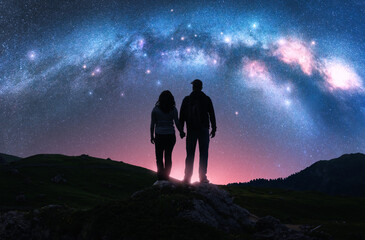 Wall Mural - Couple of hikers holding hands standing on a rocky outcrop in mountains, night pink starry sky with glowing Milky Way. Colorful landscape with silhouettes of man and woman, bright stars and milky way
