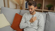 © Krakenimages.com - Young hispanic man sitting indoors with hand on chest looking concerned in a cozy apartment living room featuring a comfortable sofa and modern decor elements.
