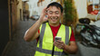 © Krakenimages.com - Young man in high-visibility vest smiling while using smartphone outdoors in urban street on sunny day, showcasing modern communication and technology use in city life
