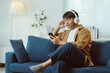 © Crystal - Relaxed young man enjoying music through wireless headphones while selecting songs on his smartphone, sitting comfortably on a sofa in his modern living room