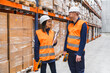 © Koldo_Studio - Two warehouse workers wearing safety vests and hard hats are discussing logistics while holding handheld devices, surrounded by shelves full of packaged goods