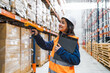 © Koldo_Studio - Smiling female warehouse worker wearing safety helmet and vest, holding clipboard and using barcode scanner for inventory management in large distribution center