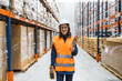 © Koldo_Studio - Smiling female warehouse worker walking through large distribution center holding barcode scanner and inventory checklist