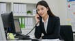 © talkative.studio - A businesswoman sits at her desk, smiling while using a landline phone.  She's professionally dressed in a suit, working in a modern office environment