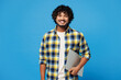 © ViDi Studio - Young smiling happy IT Indian man he wears yellow checkered shirt casual clothes hold closed laptop pc computer look camera isolated on plain blue cyan background studio portrait. Lifestyle concept.