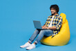 © ViDi Studio - Full body young Indian IT man wear yellow checkered shirt casual clothes sit in bag chair hold use work on laptop pc computer isolated on plain blue cyan background studio portrait. Lifestyle concept.