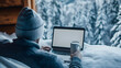 © Atirut - Person in winter attire using a laptop outdoors on a snowy landscape, holding a mug, with a backdrop of snow-covered trees.