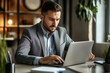© Zeljka - A man in a gray suit is seated at a desk in a modern office, focused on his laptop screen.