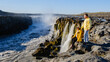 © Fokke Baarssen - Two travelers marvel at the grandeur of Dettifoss, one of Icelands most powerful waterfalls. The sun illuminates the rocky landscape, creating a breathtaking experience for nature enthusiasts.