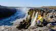 © Fokke Baarssen - Visitors sit on rocky terrain, captivated by the mighty Dettifoss waterfall in Iceland.