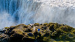 © Fokke Baarssen - Adventurers stand near a powerful Dettifoss waterfall in Iceland, surrounded by lush green moss and rocky terrain.