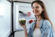 © SHOTPRIME STUDIO - Young woman enjoying a green smoothie in front of an open fridge, promoting healthy eating and a vibrant lifestyle with fresh produce