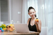 © Natthawadee - A smiling young woman in activewear holds broccoli while looking at a laptop, promoting healthy eating and digital engagement.